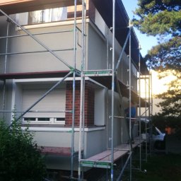 MP-BUD Usługi Budowlane - Dwupiętrowy dom w trakcie prac elewacyjnych, otoczony rusztowaniem, z widocznym fragmentem dachu z czerwonej dachówki i elementami dekoracyjnymi z czerwonej cegły.