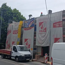 Dwóch robotników na dachu budynku w trakcie prac, zabezpieczonego siatkami ochronnymi z reklamami, na ulicy stoi samochód dostawczy z materiałami budowlanymi i żółtą rurą zsypową.