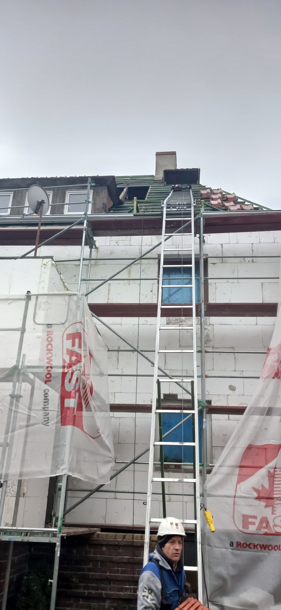 Pracownik w kasku stoi u podnóża drabiny opierającej się o budynek w trakcie prac remontowych dachu, widoczne rusztowania i materiały izolacyjne z logo firmy FAST.