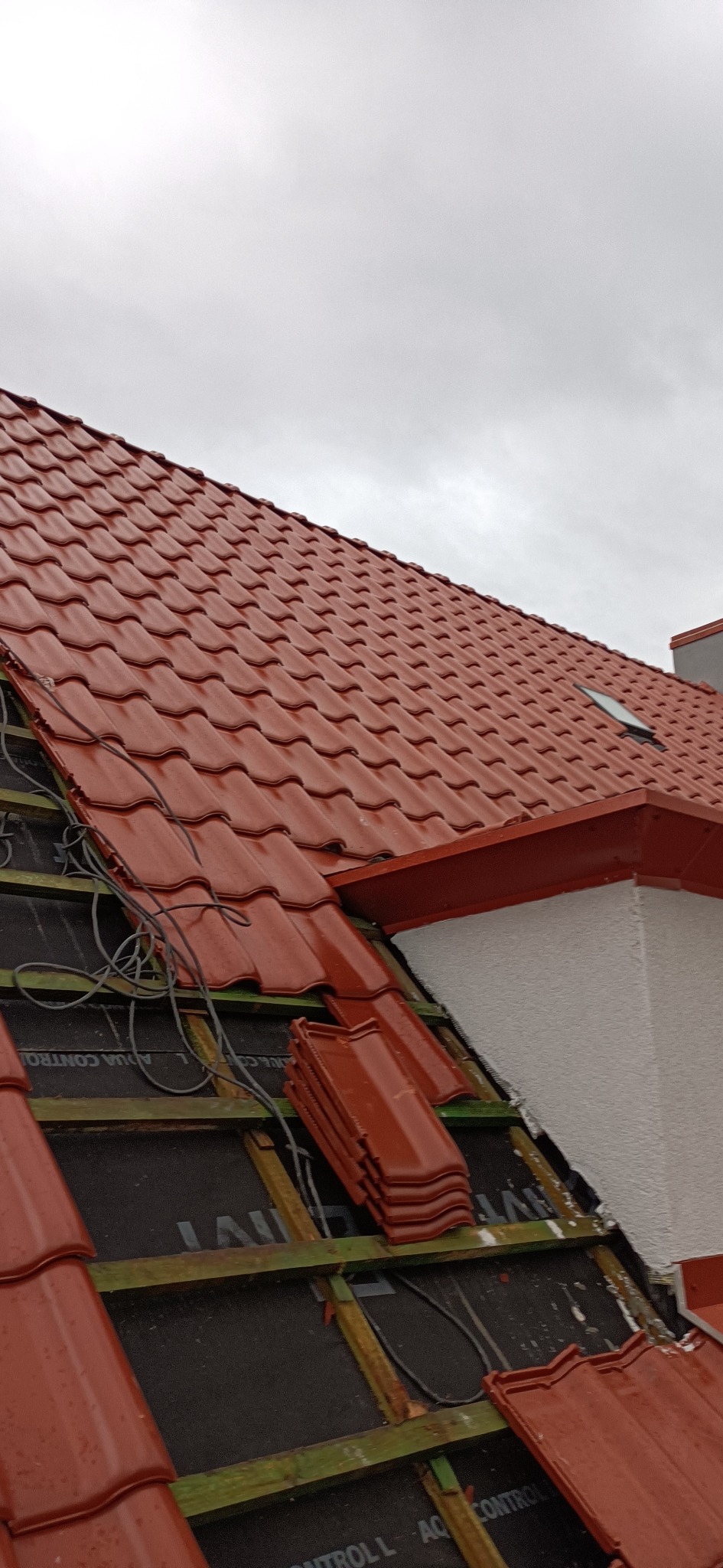 Czerwona dachówka ceramiczna układana na drewnianej konstrukcji dachu, widoczne fragmenty membrany dachowej i przewody elektryczne, pochmurne niebo w tle.