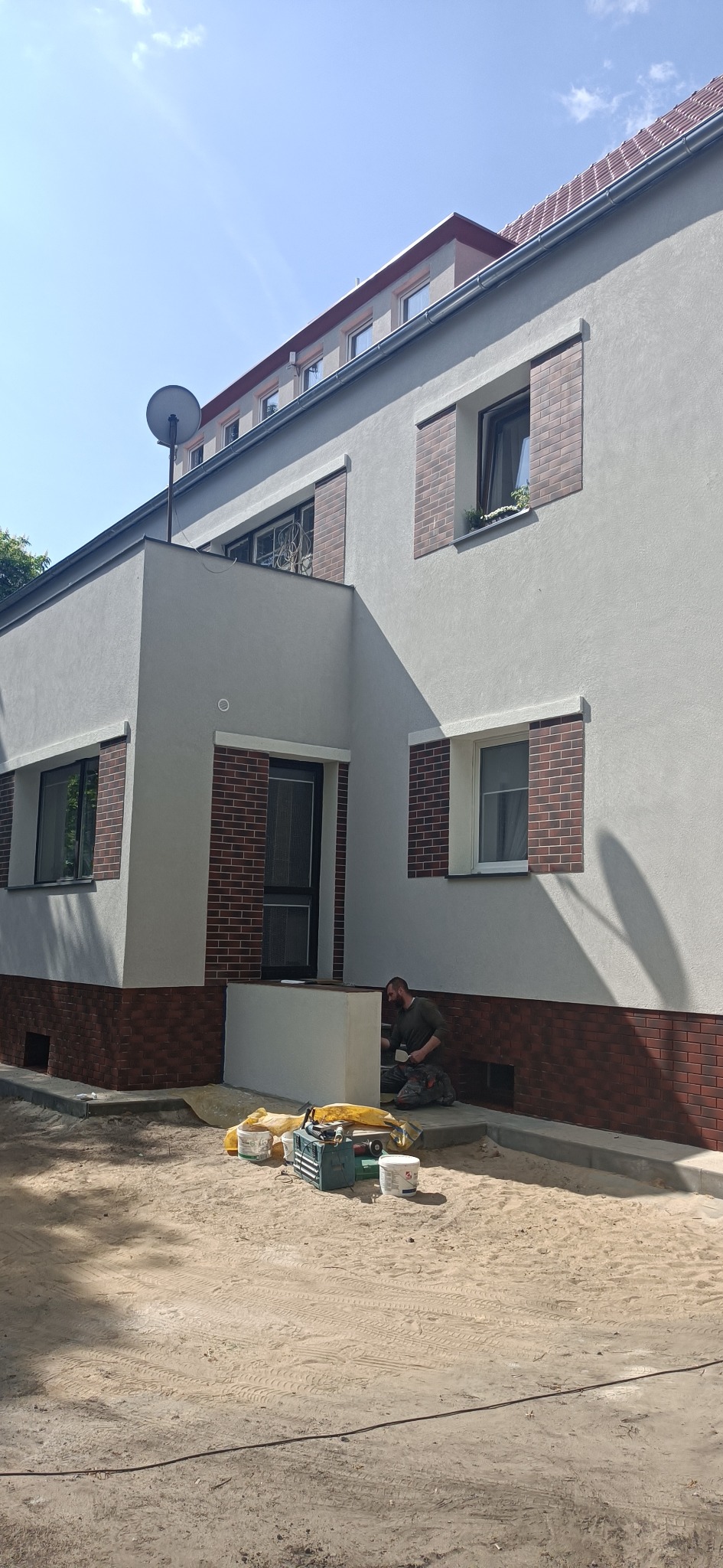 Pracownik maluje dolną część elewacji budynku, widoczne narzędzia malarskie, pędzle, wiaderka z farbą i skrzynka narzędziowa, budynek z oknami w ceglanej oprawie i anteną satelitarną na dachu.