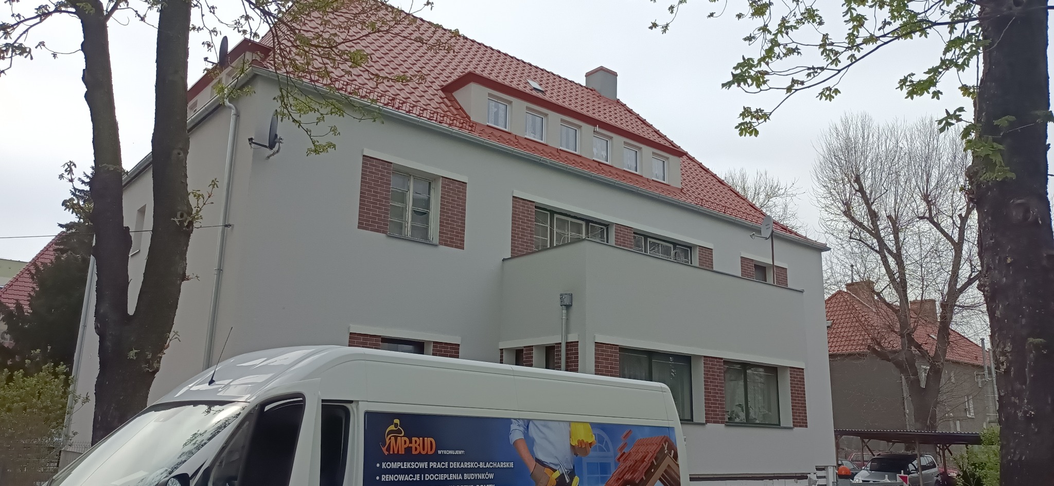 Odnowiony budynek z nową czerwoną dachówką, białą elewacją i ceglanymi obramowaniami okien, częściowo zasłonięty przez drzewa i pojazd firmy budowlanej z reklamą usług dekarskich i dociepleń.