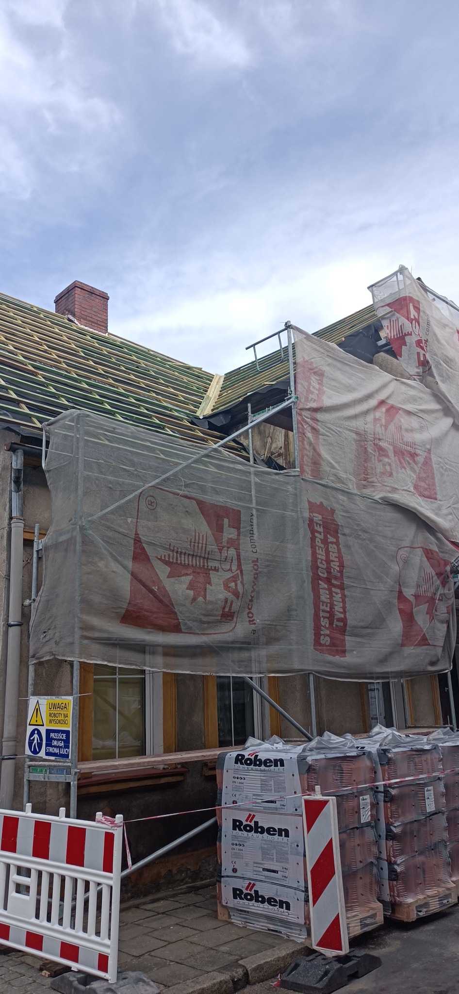 Budynek w trakcie prac dekarskich, widoczne rusztowanie z siatką ochronną, częściowo zdjęte stare pokrycie i przygotowane drewniane łaty, palety z dachówkami Roben na chodniku, znak ostrzegawczy...