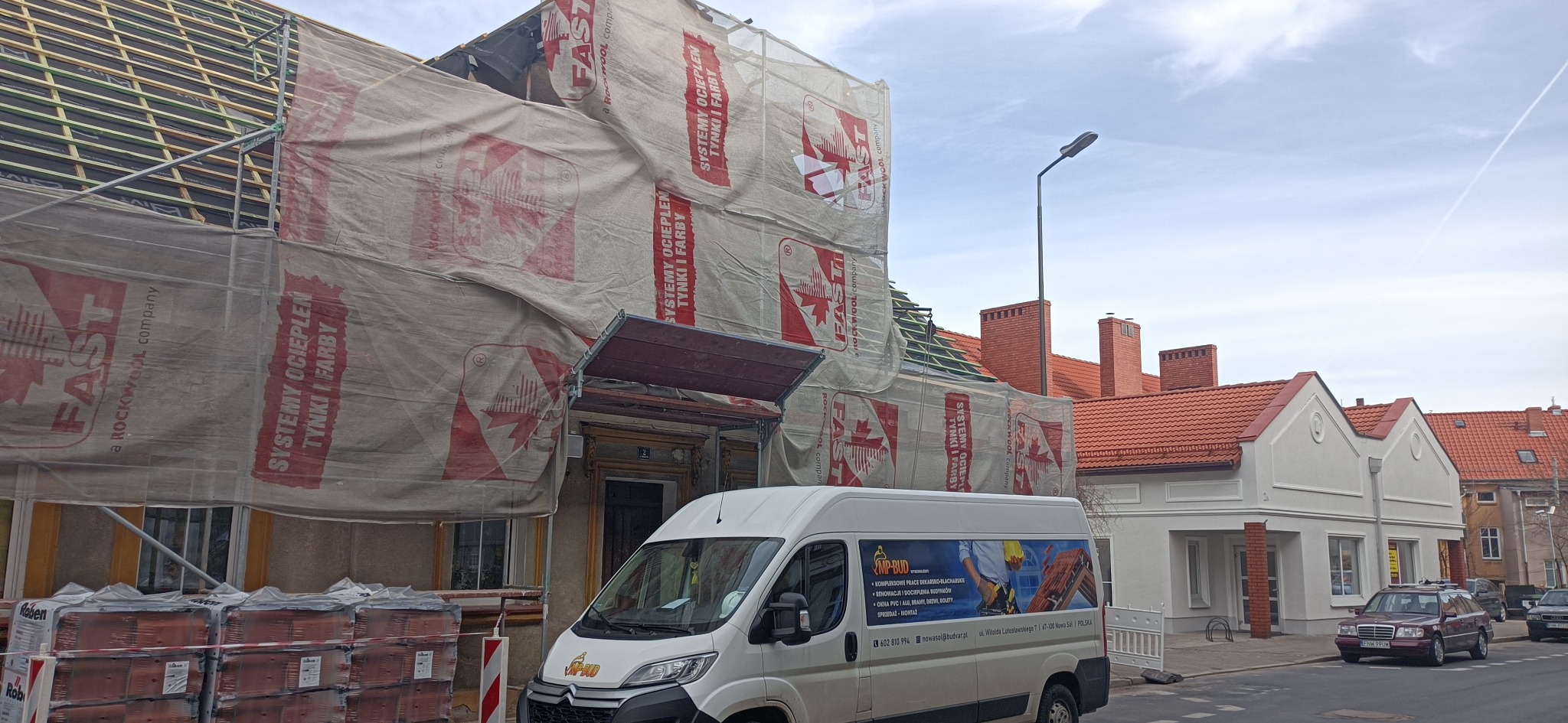 Budynek w trakcie prac dekarskich, zabezpieczony folią z logo producenta materiałów izolacyjnych, widoczne krokwie dachu, palety z dachówkami na pierwszym planie i samochód dostawczy firmy dekarskiej.