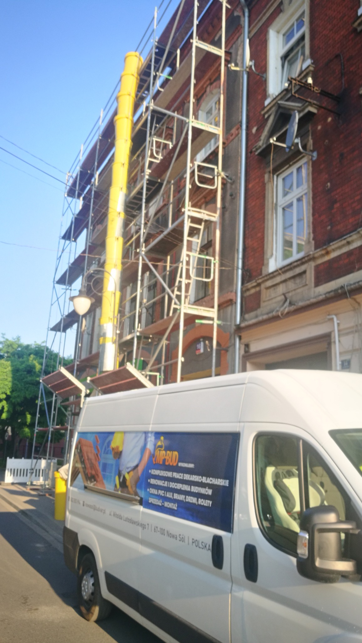 Budynek w trakcie renowacji elewacji z rusztowaniem i żółtym zsypem na gruz, na pierwszym planie samochód firmy budowlanej z reklamą usług dekarsko-blacharskich i dociepleń.