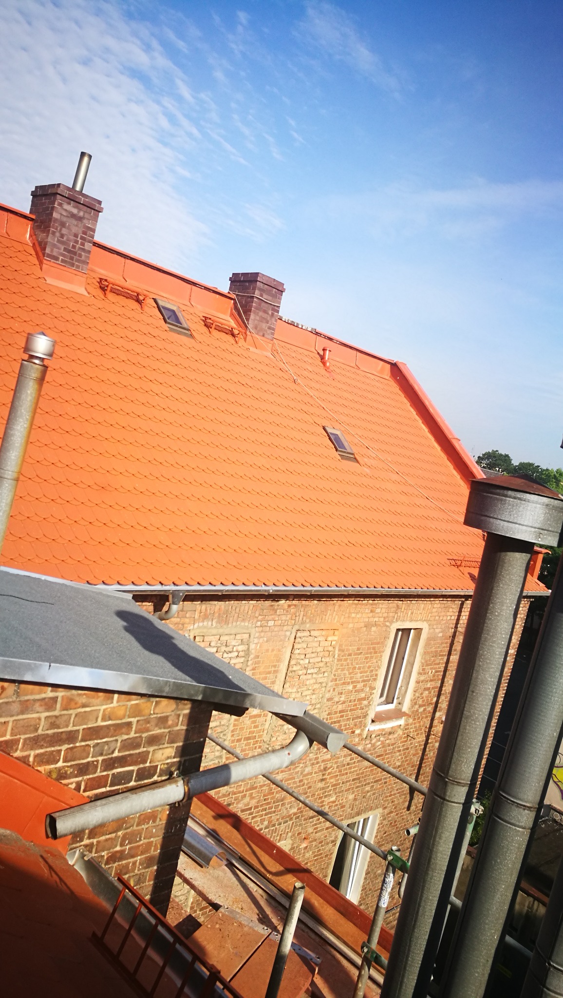 Czerwony dach z dachówki karpiówki na ceglanym budynku, widoczne kominy z cegły klinkierowej, okna dachowe, rynna i fragment rusztowania na pierwszym planie, błękitne niebo w tle.