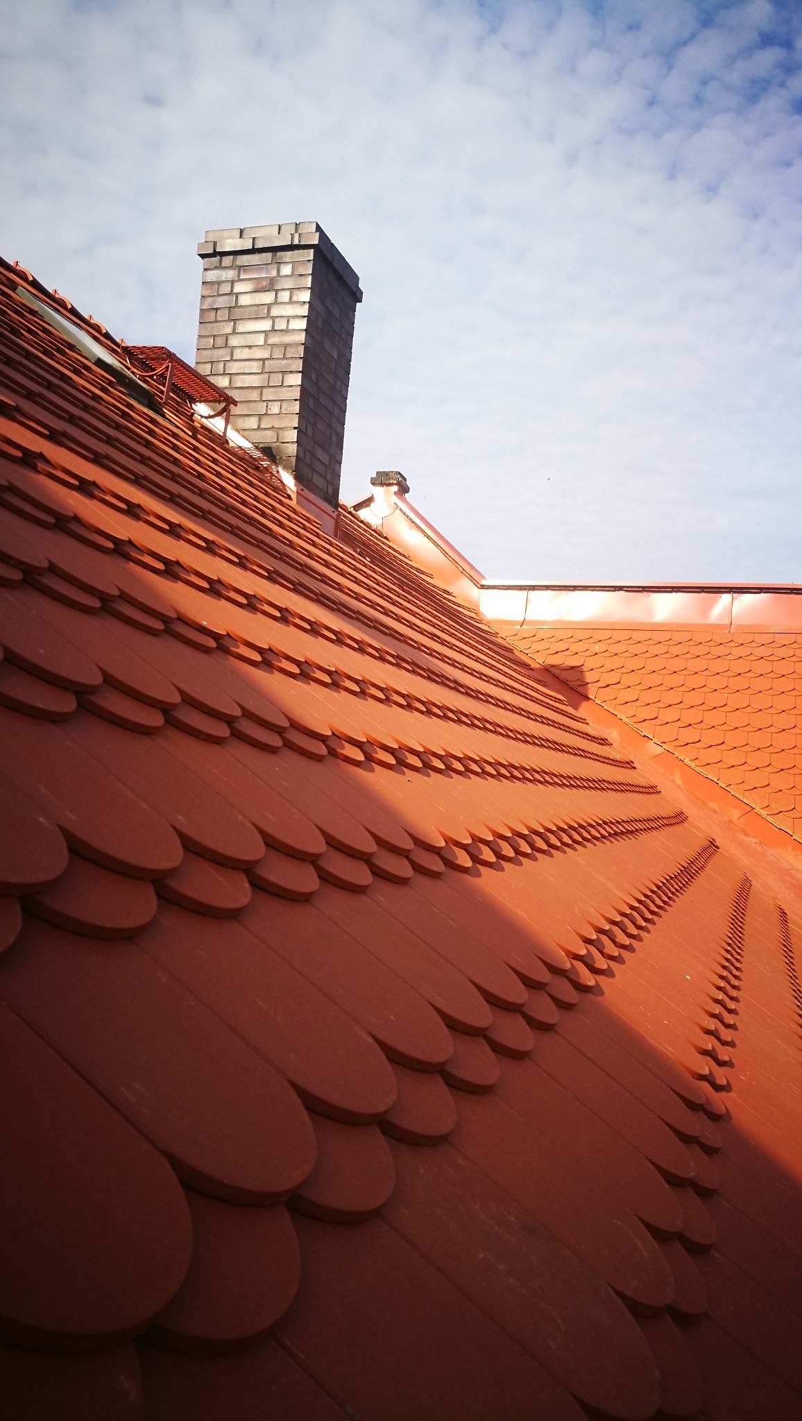 Nowy dach pokryty dachówką karpiówką w odcieniach czerwieni, z widocznym kominem z cegły i fragmentem nieba z lekkimi chmurami.