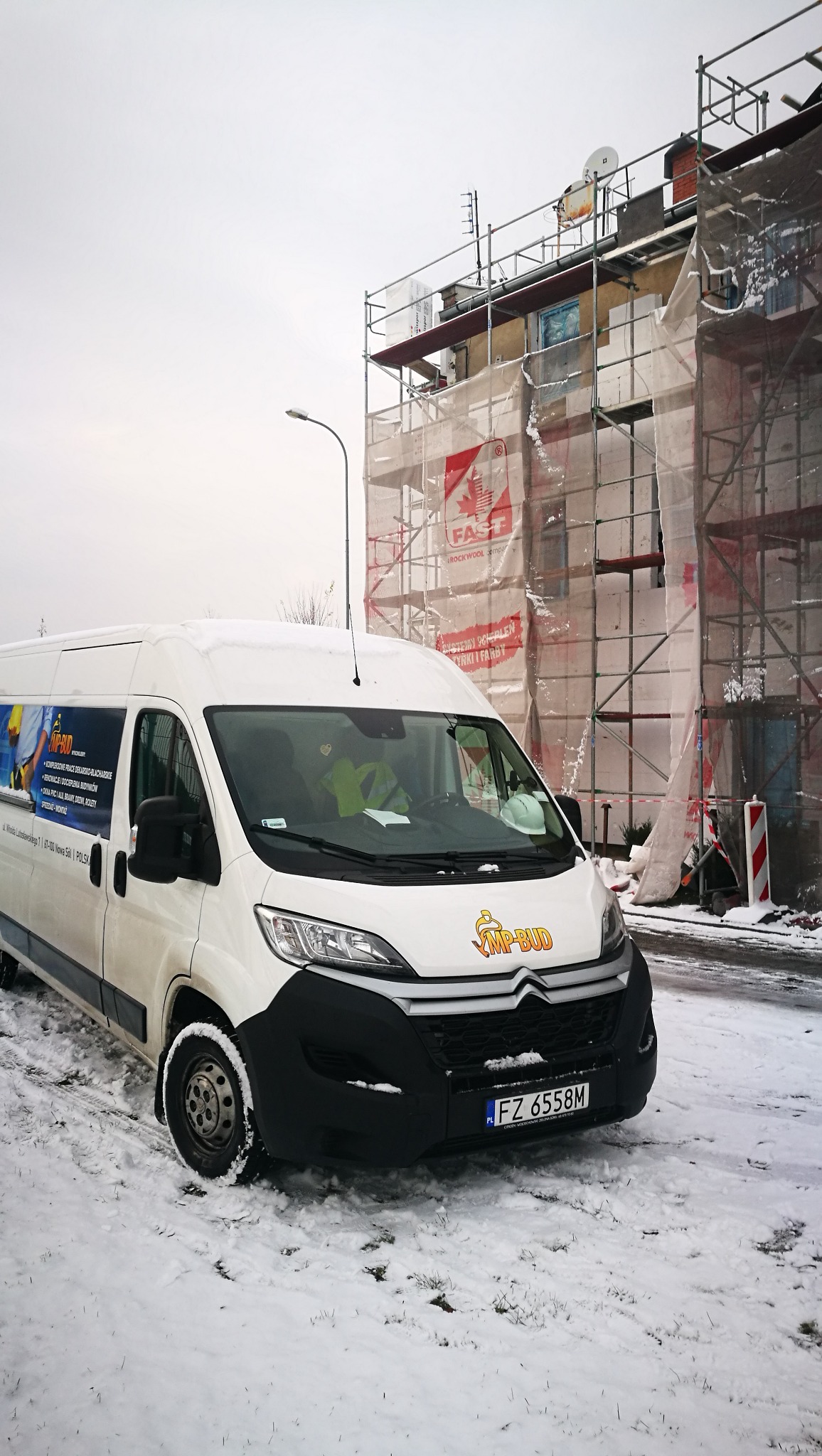 Biały bus firmy budowlanej na tle budynku w rusztowaniach, zabezpieczonego siatką podczas prac elewacyjnych w zimowej scenerii.