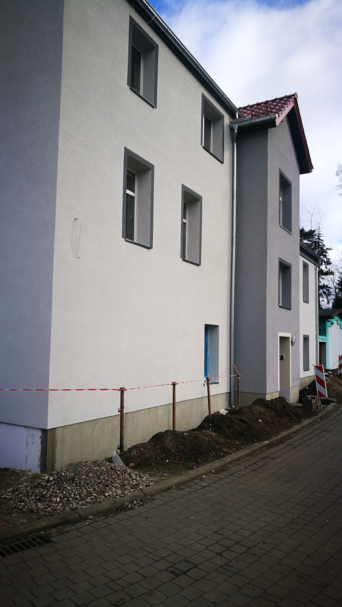 Dwukondygnacyjny budynek po ociepleniu elewacji, widoczne świeżo otynkowane ściany w odcieniach szarości i białe obramowania okien, teren wokół budynku częściowo rozkopany, zabezpieczony taśmą.