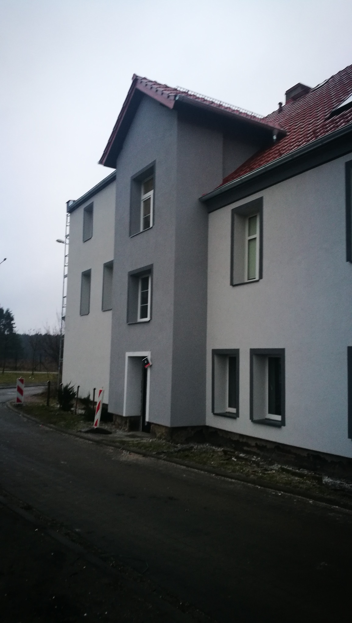Dwukondygnacyjny budynek po ociepleniu i pomalowaniu elewacji na odcienie szarości, z czerwoną dachówką, widok z poziomu ulicy w pochmurny dzień.