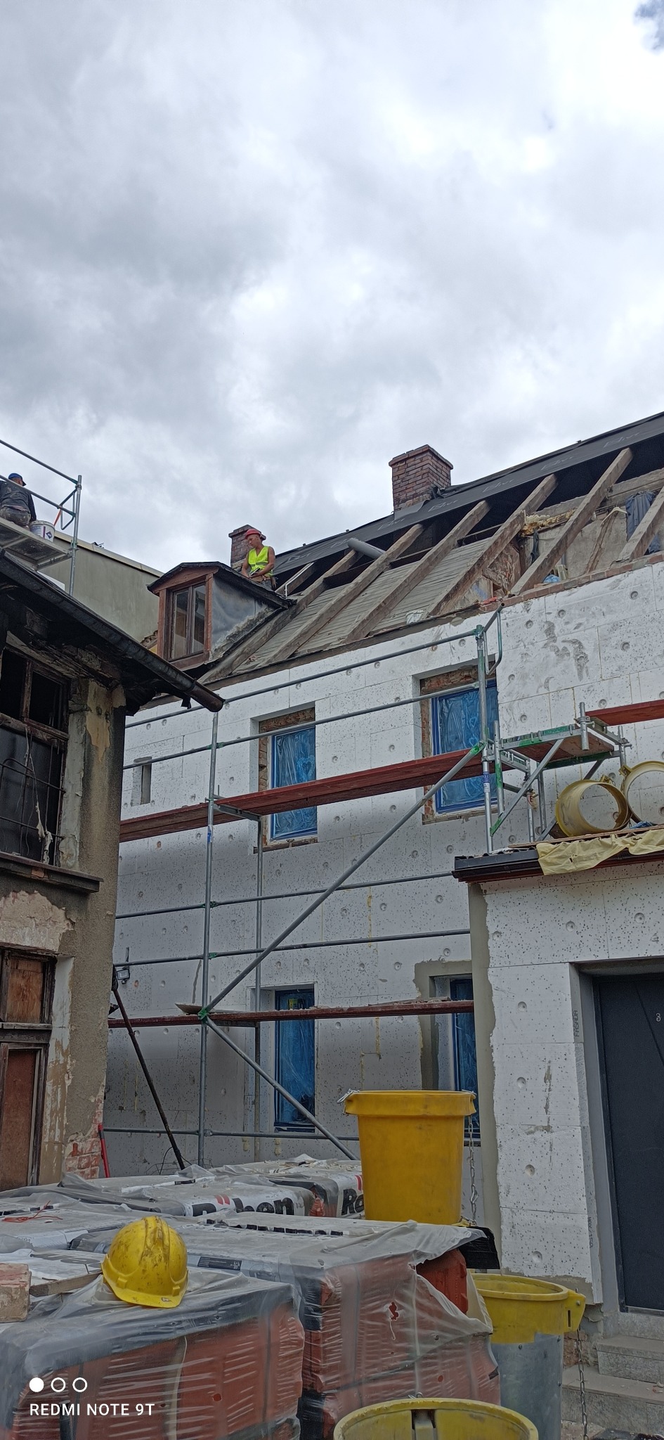 Budynek w trakcie prac remontowych: ocieplanie elewacji styropianem, częściowo zdjęte pokrycie dachowe, widoczne rusztowania, pracownicy na dachu, okna zabezpieczone folią.