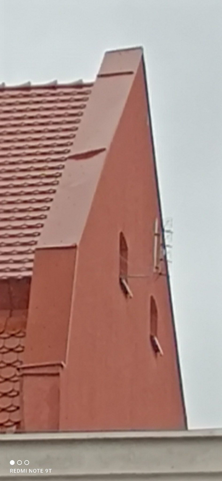 Czerwony budynek z dwoma wąskimi oknami i anteną na ścianie, widoczny fragment dachu z dachówką karpiówką.