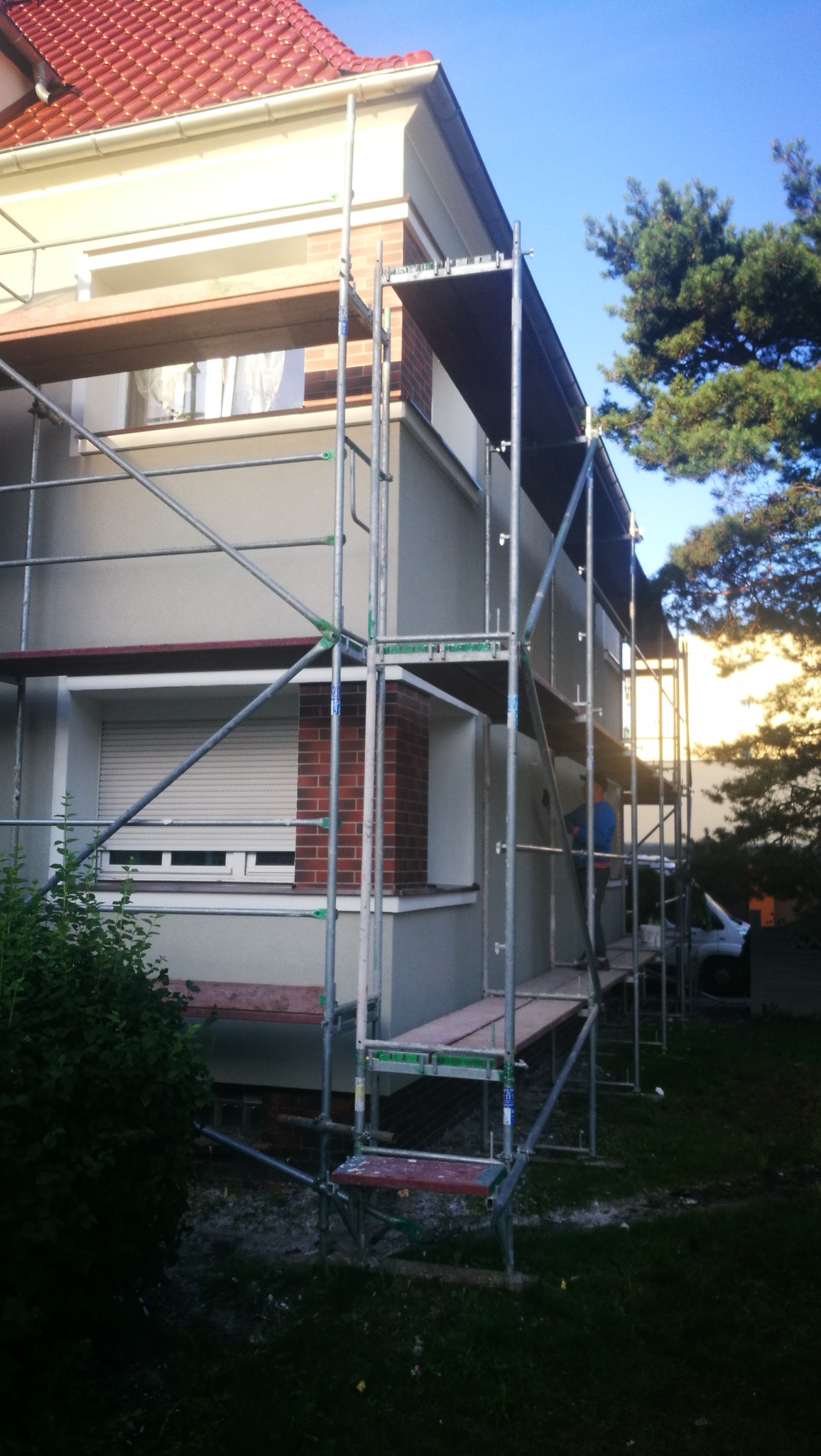 Dwupiętrowy dom w trakcie prac elewacyjnych, otoczony rusztowaniem, z widocznym fragmentem dachu z czerwonej dachówki i elementami dekoracyjnymi z czerwonej cegły.