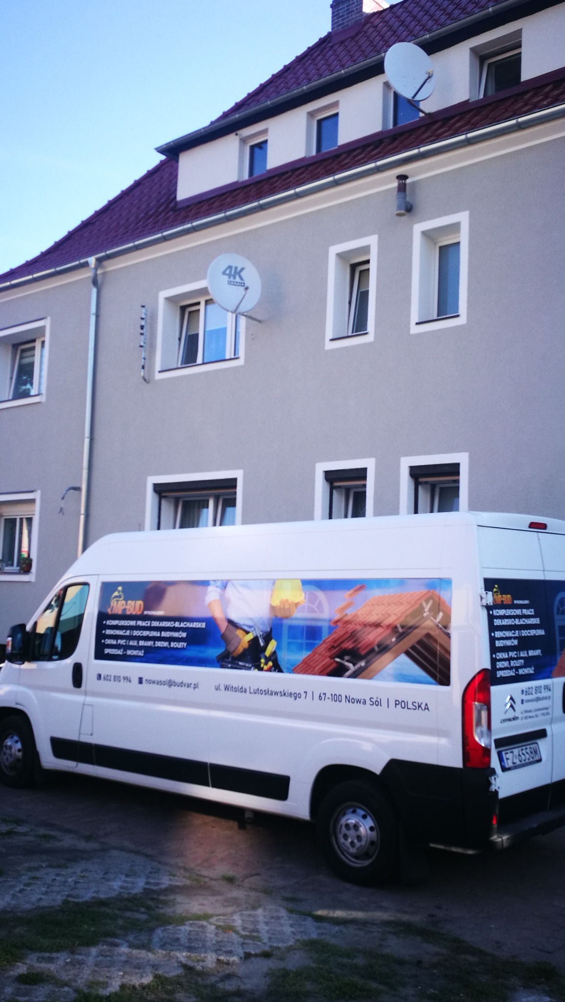 Budynek mieszkalny z nowym, bordowym dachem widziany z perspektywy ulicy, z zaparkowanym białym samochodem dostawczym firmy dekarskiej z reklamą usług na boku.
