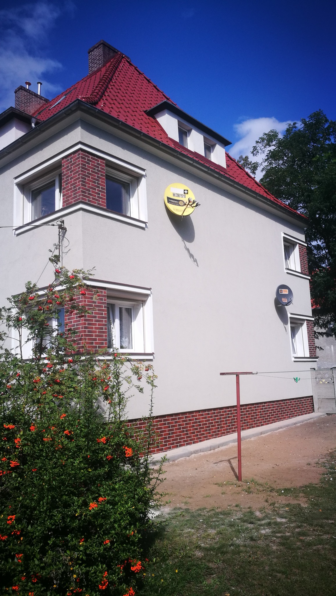 Odnowiony budynek z czerwoną dachówką, elewacją w kolorze złamanej bieli i ceglanymi detalami wokół okien, antena satelitarna na ścianie, krzew jarzębiny na pierwszym planie.
