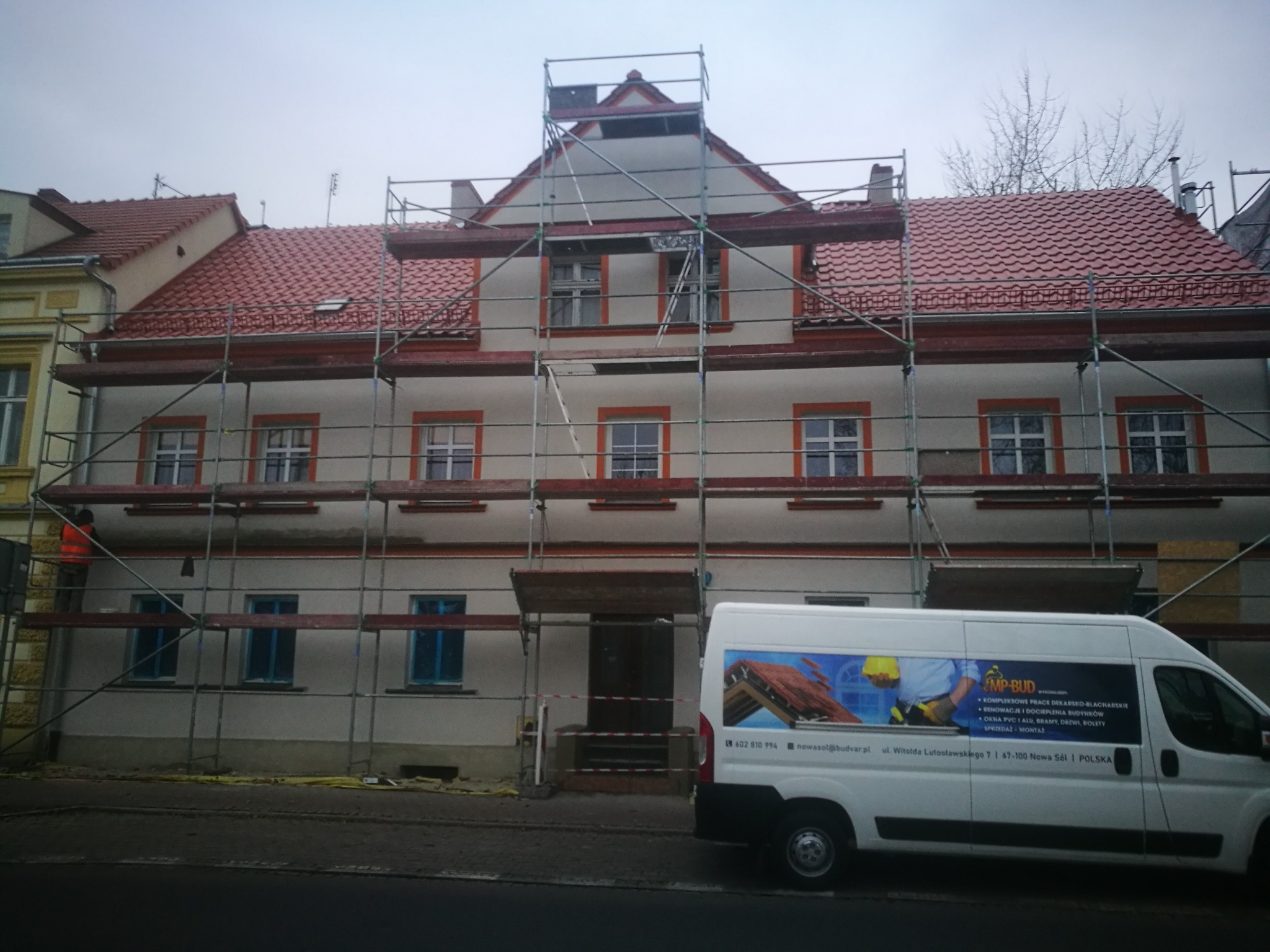 Budynek dwupiętrowy w trakcie prac remontowych, otoczony rusztowaniami, z częściowo wymienionym, czerwonym dachem i oknami zabezpieczonymi folią, na pierwszym planie samochód dostawczy z reklamą...