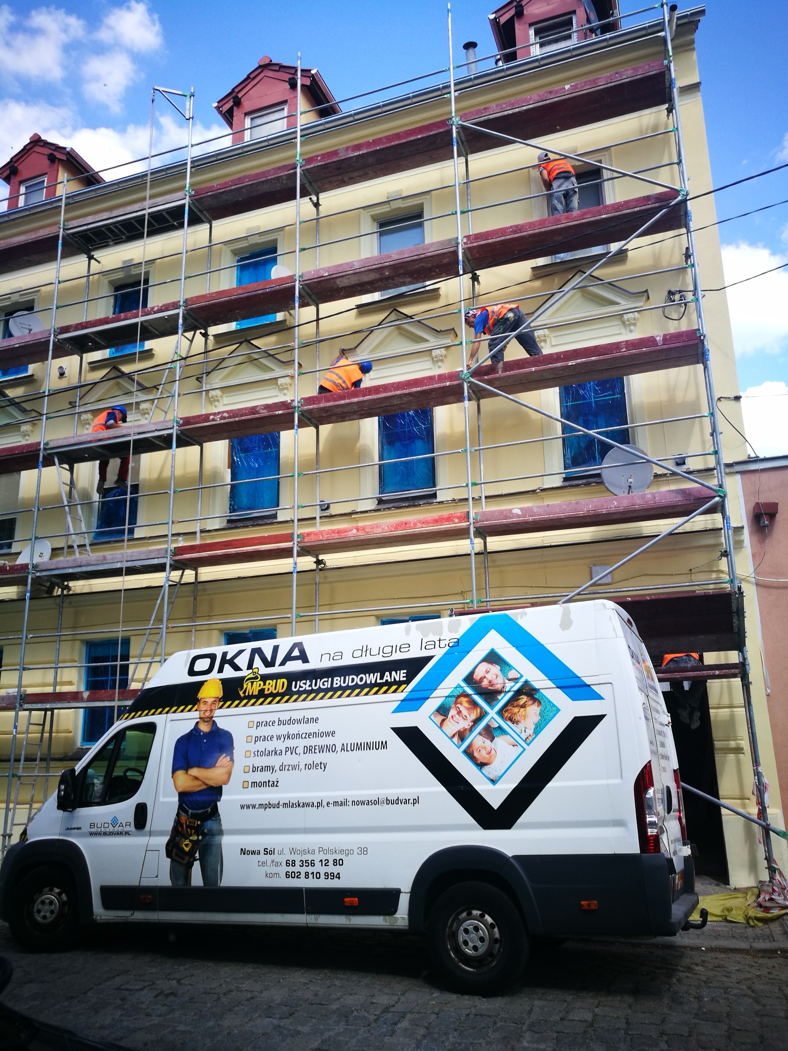 Pracownicy na rusztowaniu podczas prac elewacyjnych na budynku z żółtą fasadą, okna zabezpieczone folią, na pierwszym planie samochód dostawczy z reklamą firmy budowlanej.