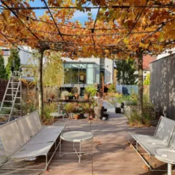 Nowoczesny taras z drewnianą podłogą, meblami ogrodowymi w odcieniach beżu i zadaszeniem z gałęzi drzewa o jesiennych liściach.