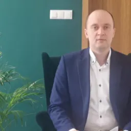 Mężczyzna w marynarce siedzi w fotelu na tle zielonej ściany z włącznikiem światła i drewnianych drzwi, obok stoi roślina doniczkowa.