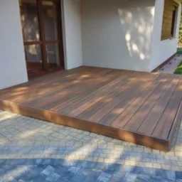 Nowoczesny taras kompozytowy w odcieniach brązu, przylegający do jasnego budynku z brązowymi oknami, wykończony kostką brukową w szaro-niebieskich tonacjach.