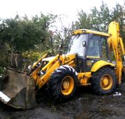 Żółta koparko-ładowarka JCB z opuszczoną łyżką na ziemi, widoczne drzewa w tle.