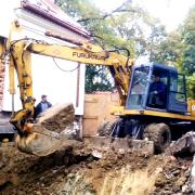 Żółta koparka Furukawa z operatorem w trakcie wykopu fundamentów, widoczny fragment budynku w tle.