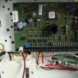 Centrala alarmowa Satel CA-6 z widocznymi podłączeniami kablowymi wewnątrz białej obudowy. Widoczne elementy elektroniczne, złącza śrubowe i przewody różnych kolorów.