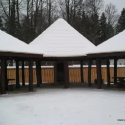 Zadaszona, drewniana altana z miejscami do siedzenia pokryta śniegiem, otoczona zimowym krajobrazem.