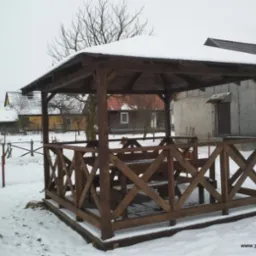 Drewniana altana ogrodowa z zadaszeniem, pokryta śniegiem, otoczona śnieżnym krajobrazem z widocznymi budynkami w tle.