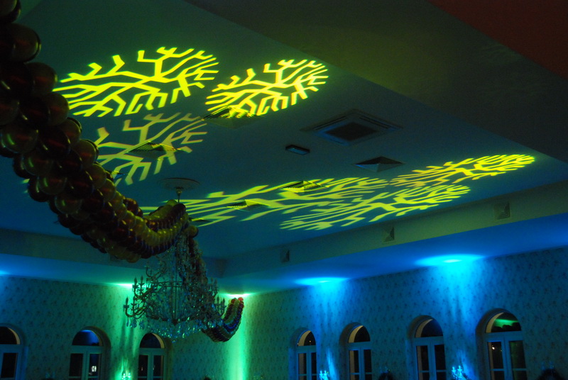 Sala balowa z dekoracją świetlną w postaci żółtych wzorów drzew na suficie, kryształowy żyrandol i girlanda z balonów, oświetlenie ścian w kolorze niebieskim i zielonym.