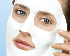 Twarz kobiety z nałożoną białą maską kosmetyczną; widoczne wycięcia na oczy i usta, delikatny dotyk palca do skóry.