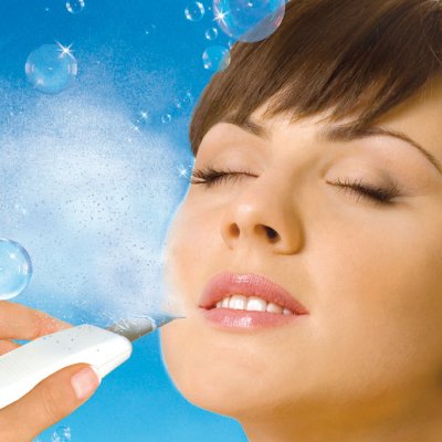 Kobieta z zamkniętymi oczami podczas zabiegu mikrodermabrazji diamentowej, widoczna dysza urządzenia w pobliżu policzka, błękitne tło z bąbelkami.