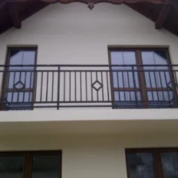 Czarna, metalowa balustrada balkonowa z motywem rombu, widok z dołu na jasną elewację budynku i stolarkę okienną.