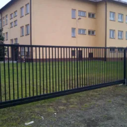 Czarna, przesuwna brama wjazdowa z pionowych prętów na tle budynku z żółtą elewacją i zielonym trawnikiem.