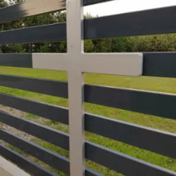 Nowoczesne ogrodzenie panelowe z metalowymi poprzeczkami w kolorze grafitowym i ozdobnym elementem w kształcie krzyża, widok z bliska na tle zielonego trawnika i drzew.