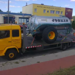 Żółta laweta z napisem 'Pomoc Drogowa 24h Transport Maszyn' przewozi srebrny zbiornik z napisem 'JOBKIN Modulo 2' na tle budynku z napisem 'HOTEL RESTAURACJA PIZZERIA'.