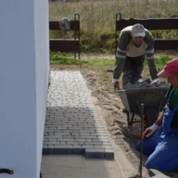 Układanie kostki brukowej na ścieżce obok budynku. Dwóch mężczyzn w strojach roboczych i wózek z kostką. Częściowo ułożona nawierzchnia.