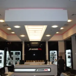 Wnętrze sklepu z elektroniką X-Kom.pl z oświetleniem sufitowym, czarnymi ścianami i ekspozycją laptopów.