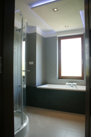 Nowoczesna łazienka z kabiną prysznicową z zaokrąglonym szkłem, podświetlanym sufitem i oknem z drewnianą ramą nad wanną.