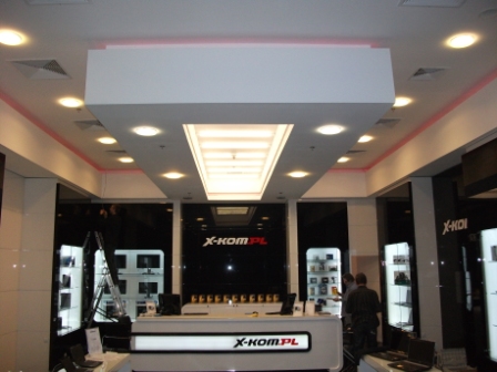 Wnętrze sklepu z elektroniką X-Kom.pl z oświetleniem sufitowym, czarnymi ścianami i ekspozycją laptopów.
