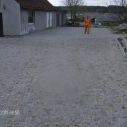 Ułożona szara kostka granitowa na podwórku z pracownikiem w pomarańczowym kombinezonie w tle, widoczne fragmenty budynku i schodów w trakcie budowy.