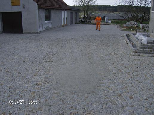 Ułożona szara kostka granitowa na podwórku z pracownikiem w pomarańczowym kombinezonie w tle, widoczne fragmenty budynku i schodów w trakcie budowy.
