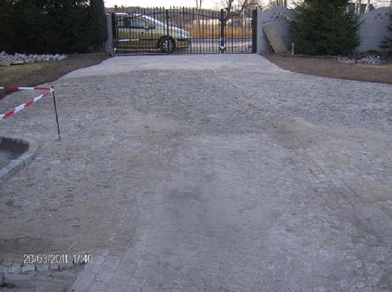 Niedokończona droga dojazdowa z kostki, częściowo zasypana piaskiem, z widoczną bramą wjazdową i samochodem w tle, zabezpieczona taśmą ostrzegawczą.