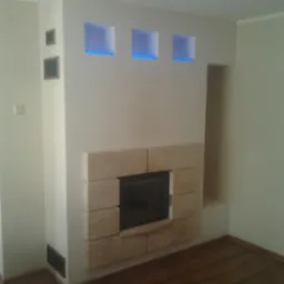 Nowoczesny kominek z obudową z jasnego kamienia, wnęki z niebieskim podświetleniem, drewniana podłoga, minimalistyczny wystrój wnętrza.