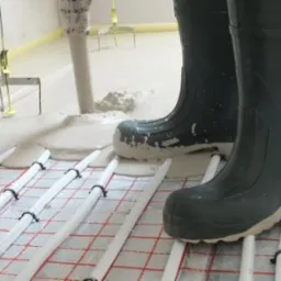 Wylewanie płynnej masy na ogrzewanie podłogowe, widoczne kalosze robotnika i narzędzia do rozprowadzania wylewki.