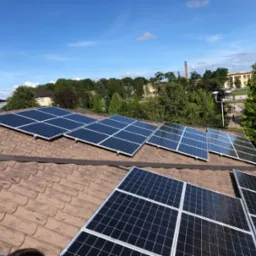 Panele fotowoltaiczne zamontowane na brązowym dachu z dachówki ceramicznej, widok z góry na tle zieleni i błękitnego nieba.