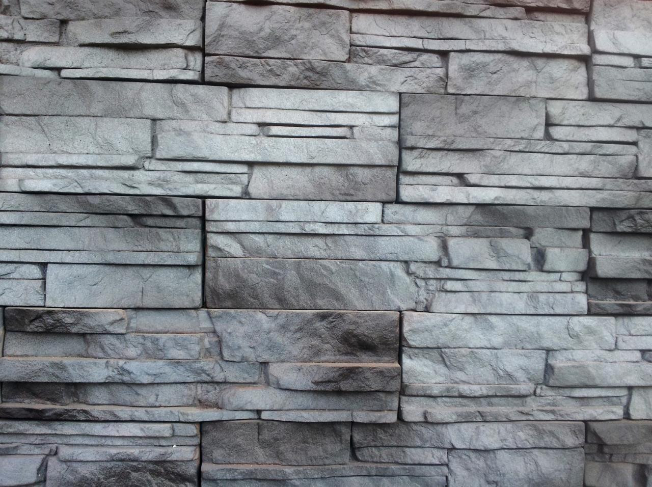 Szara ściana wyłożona kamieniem dekoracyjnym o nieregularnych kształtach i fakturze, imitującą naturalny kamień.