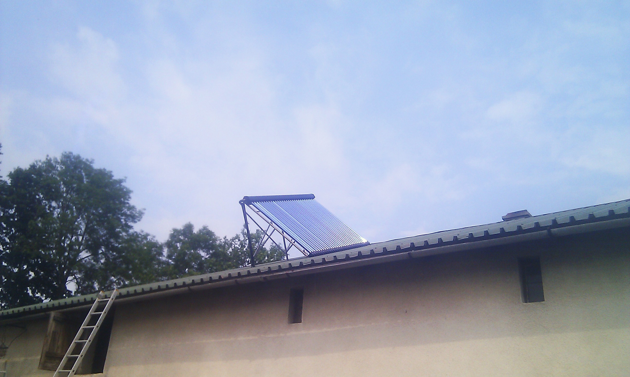 Kolektor słoneczny rurowy zamontowany na dachu budynku gospodarczego, widoczna drabina oparta o ścianę i fragment drzewa w tle.