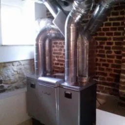 Srebrne, metalowe rury systemu wentylacyjnego połączone z centralą w pomieszczeniu z ceglaną ścianą i oknem.