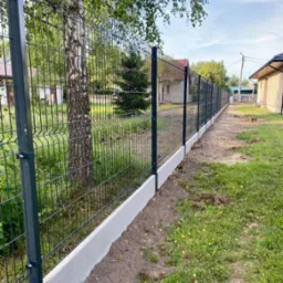 Długi, ciemny, metalowy płot panelowy z podmurówką betonową, biegnący wzdłuż trawnika i drzewa brzozy, z domami w tle.